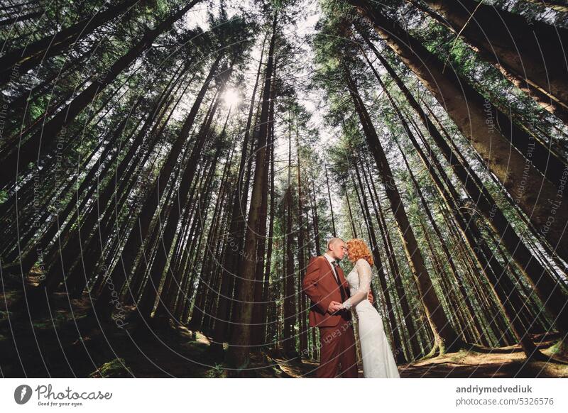 Happy stylish couple newlyweds in the green forest on summer day. bride in long white dress and groom in red suit are hugging. wedding day happy park nature