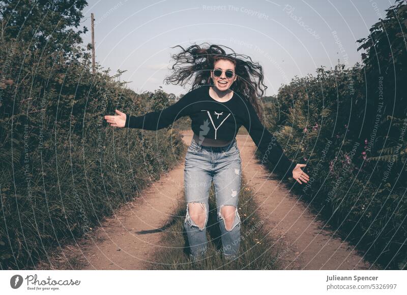 Happy girl arms outstretched wearing sunglasses on dirt track shades muted tone black jumper holes in jeans faded smiling hair flying redhead young adult fun