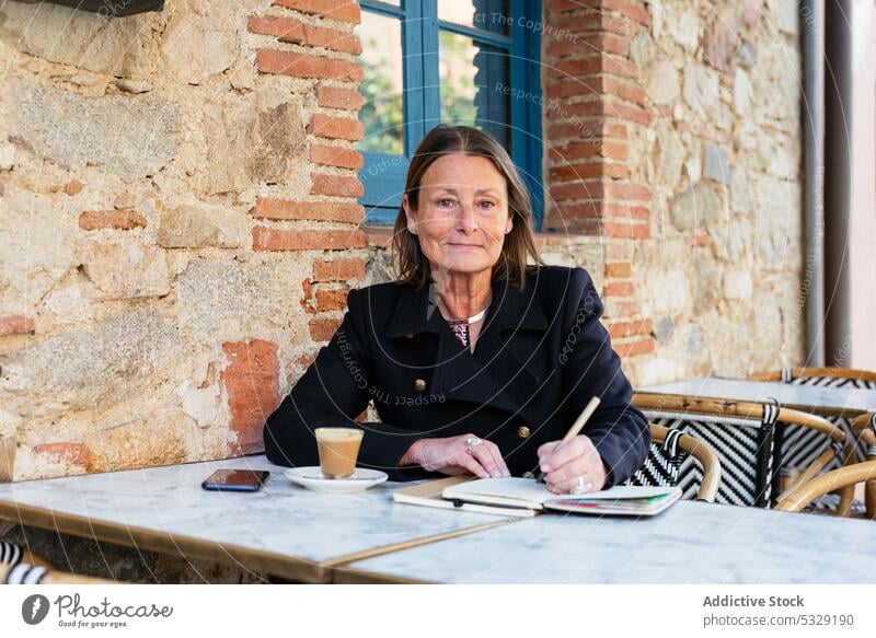 Cheerful woman with notebook sitting at table write take note cafe positive smile planner happy cheerful coffee cup female work street middle age mature glad