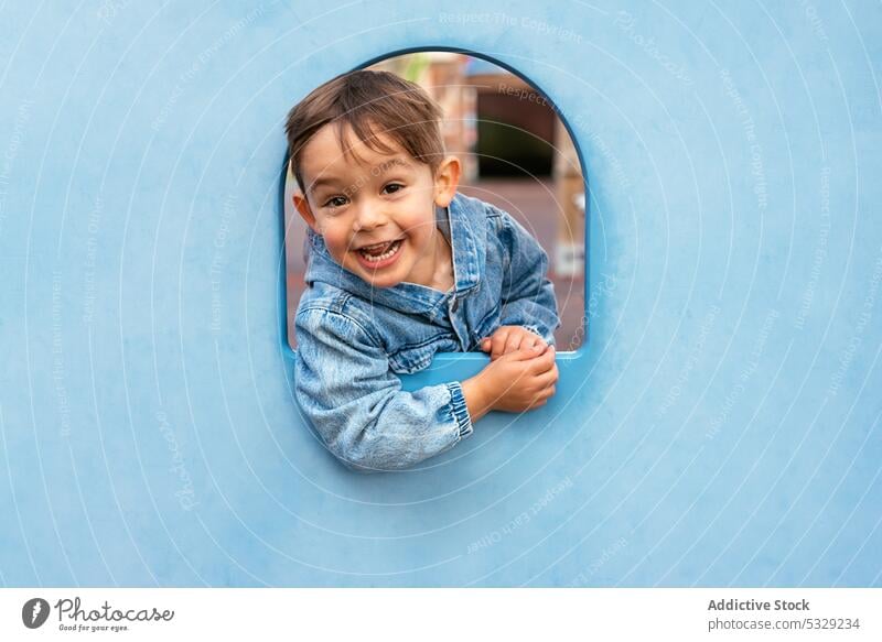 Happy little kid looking out from blue wall boy play peek playground child happy cheerful joy smile playful window childhood fun lifestyle positive casual