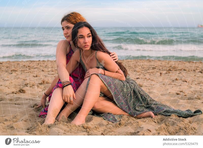 Pensive girlfriends sitting on sandy beach women thoughtful dreamy sea hug embrace tender summer ocean female shore pensive young resort wave friendship