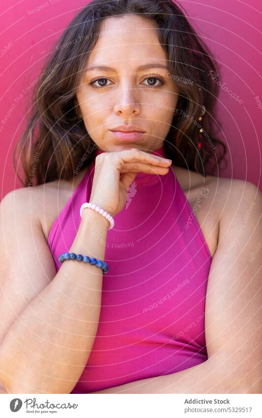 Serious woman in pink top pensive thoughtful appearance portrait calm touch chin bright vivid long hair female brunette wall dreamy young dark hair daylight