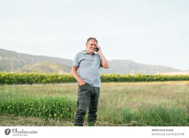 Happy mature man talking on smartphone in countryside in sunlight farmer smile hill field positive nature agriculture speak male casual communicate happy mobile