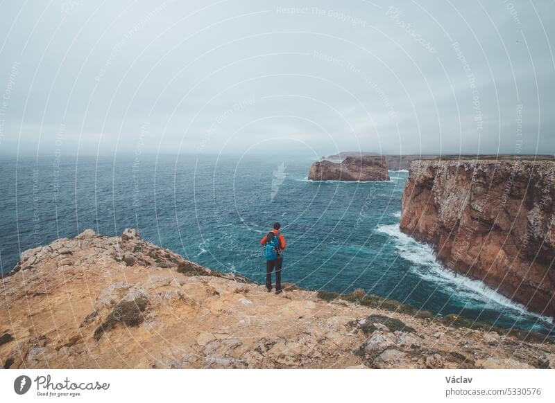 Black-haired adventurer stands at the end of Cape Cabo de Sao Vicente in the southwest of Portugal in the Algarve region. Man is enjoying his freedom. Wandering of Fisherman Trail