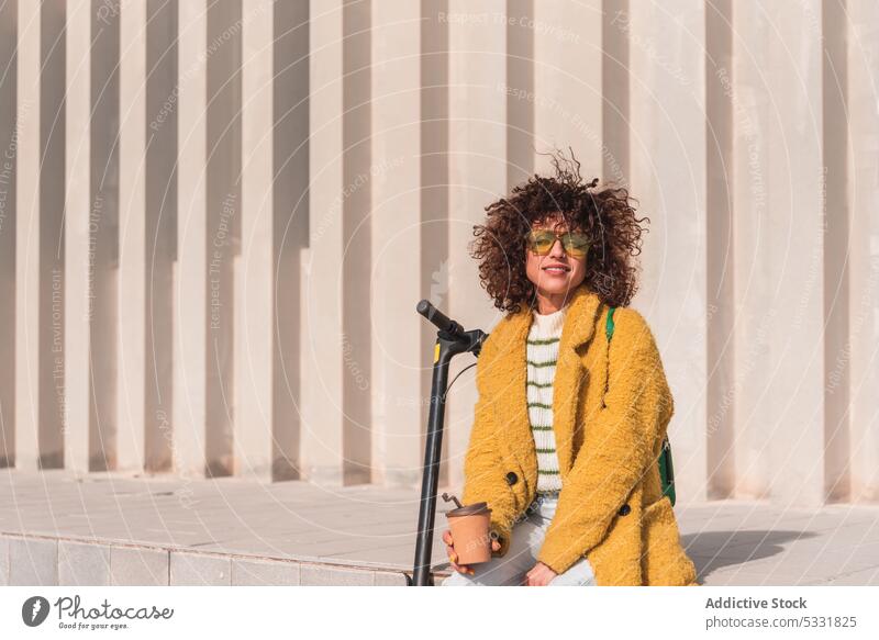 Calm woman sitting near electric scooter on street coffee takeaway trendy urban style ledge modern hipster female young transport curly hair to go city