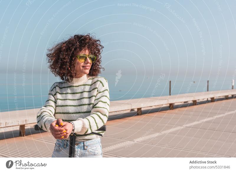 Smiling woman standing on electric scooter on embankment smile happy casual urban cheerful style curly hair glasses vehicle modern city female joy transport