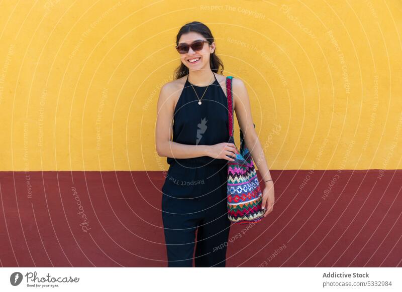 Smiling woman with handbag standing by colorful background sunglasses fashion wall positive style cheerful smile trendy bright outfit black dress apparel model