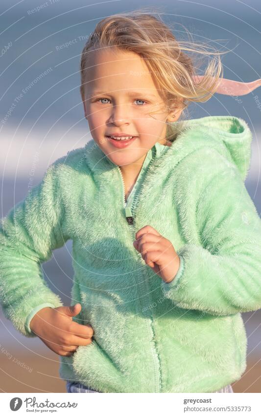 A 5-year-old girl in a green jacket is running child childhood blonde 5 years old beach sea sand ocean coast vertical young eyes blue holiday lifestyle sky