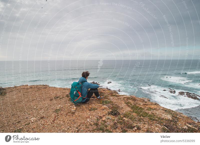 Young man sits above the famous Praia da Carreagem beach in southwest Portugal, near the town of Aljezur in the Odemira region. Wandering along the Fisherman Trail