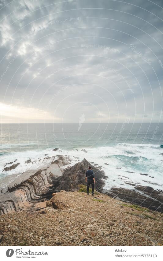 Hiker stands on top of a cliff and watches the Atlantic Ocean. Praia da Amoreira near the town of Aljezur in the Odemira Region of southwestern Portugal. Wandering along the Fisherman Trail