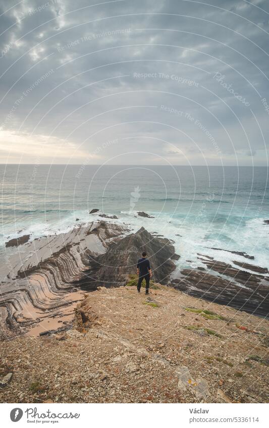 Young pilgrim stands on top of a cliff and watches the Atlantic Ocean. Praia da Amoreira near the town of Aljezur in the Odemira Region of southwestern Portugal. Wandering along the Fisherman Trail