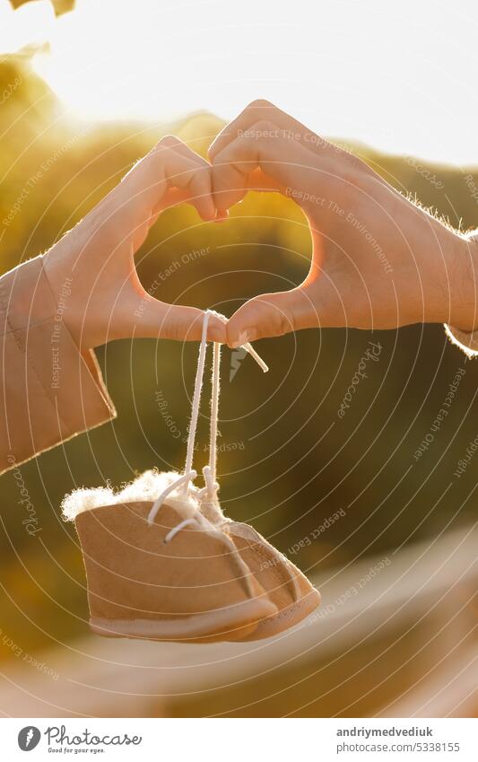 Man and pregnant wife are doing heart gesture with hands and holding warm baby shoes on nature autumn background. Future parents waiting for the baby. The concept of Mother's Day and Women's Day.