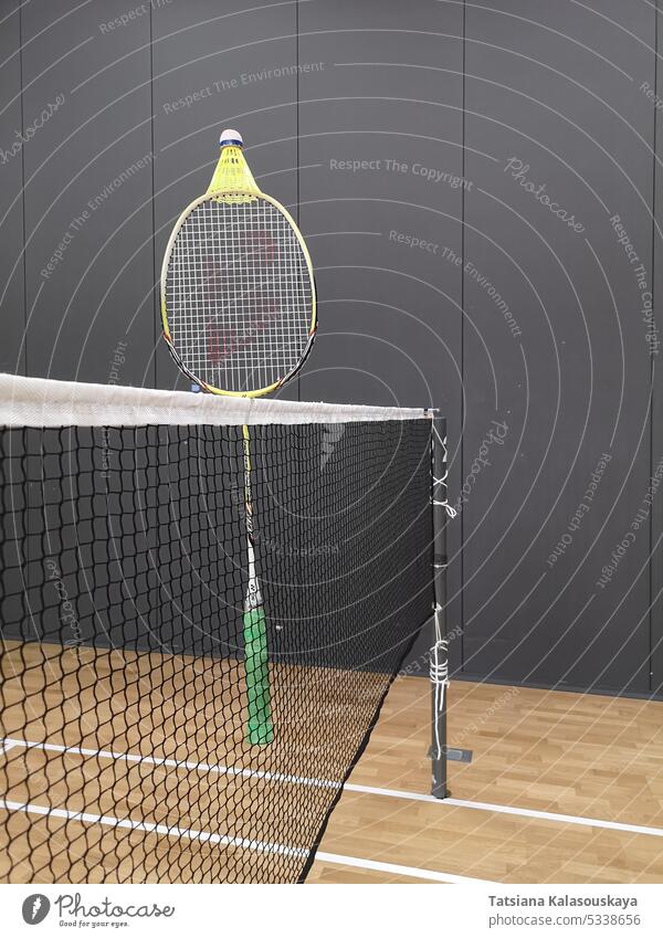 A badminton racket hangs on a badminton net, with a yellow plastic shuttlecock on top of it on an indoor badminton court. sports recreation game equipment