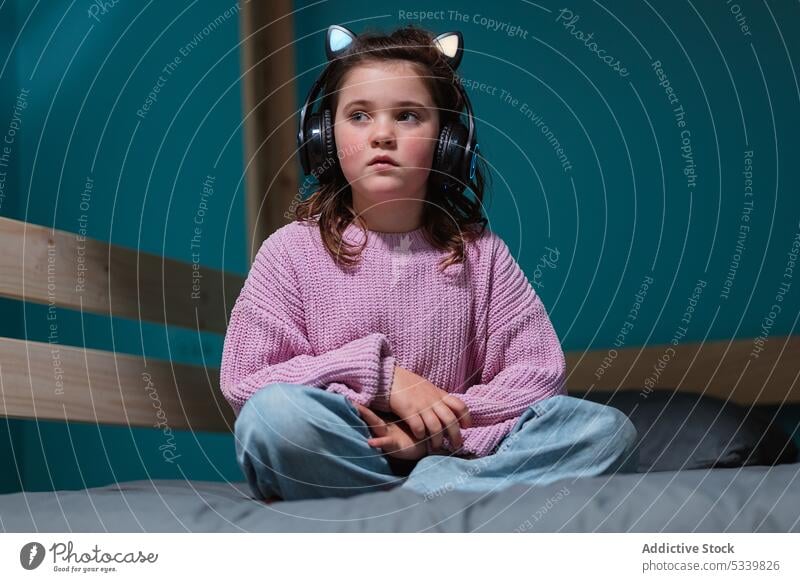 Calm child in headphones sitting on bed kid music listen legs crossed adorable thoughtful bedroom gadget little home device comfort at home calm cozy inside