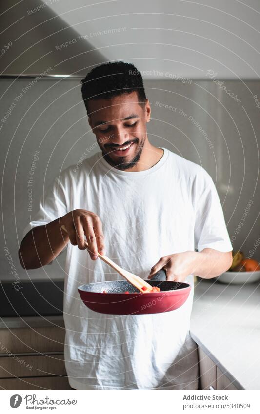 Content ethnic man frying tomatoes on pan in kitchen cook prepare mix food ingredient spatula young stir positive process casual culinary lunch smile stove