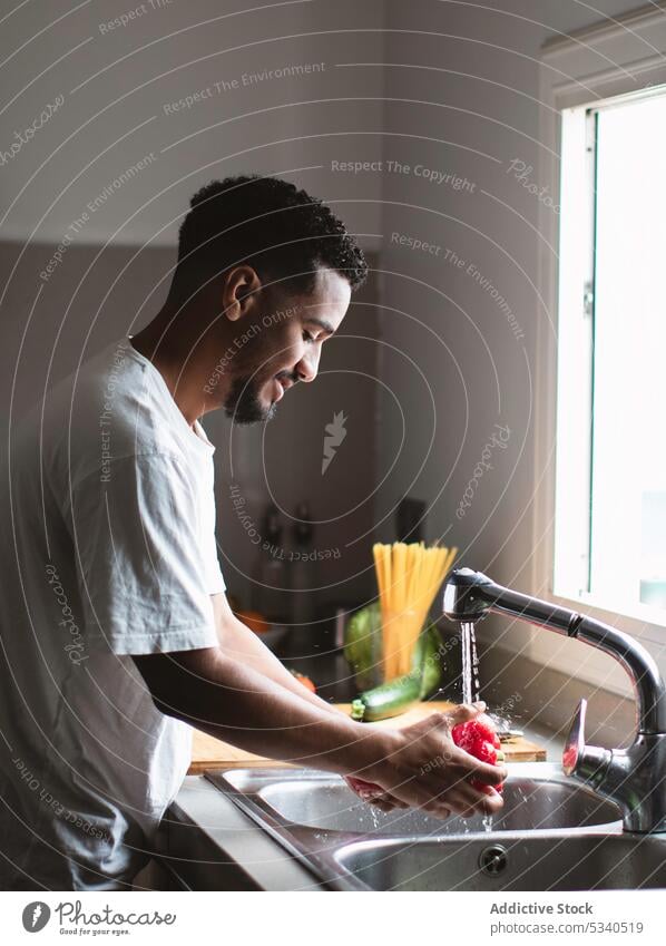 Ethnic man washing bell pepper during cooking preparation kitchen sink water prepare vegetarian fresh healthy food casual focus tap housework young vegetable