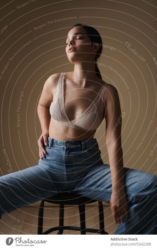 Graceful woman sitting on chair in light studio style lingerie portrait model thoughtful sensual stool jeans fashion eyes closed underwear figure female