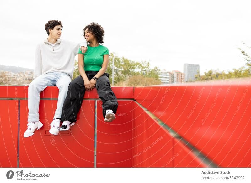 Happy diverse couple sitting on red fence man rest confident sunlight calm cloudless summer style relationship relax multiracial multiethnic boyfriend