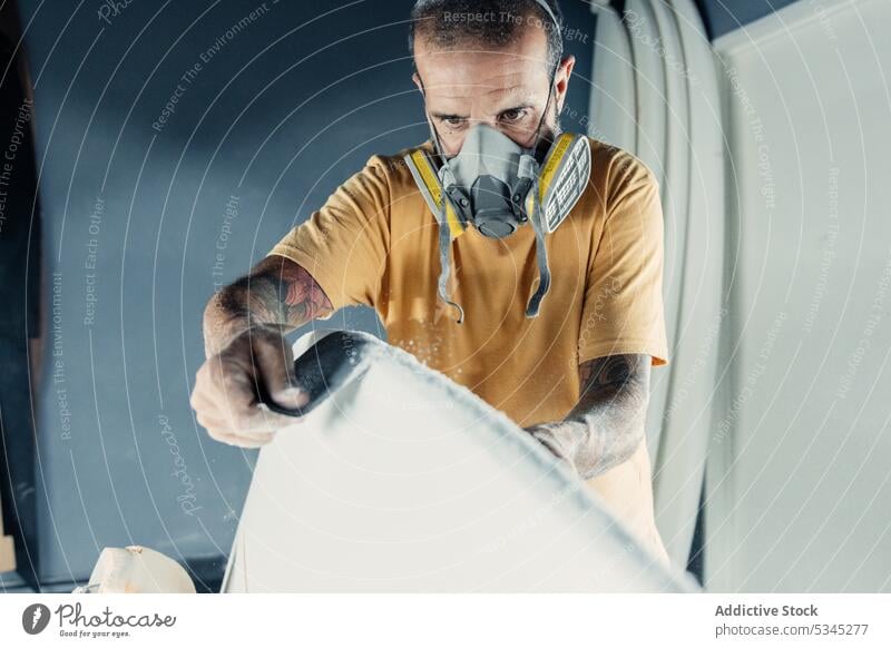 Focused man using sandpaper on surfboard polish work workshop professional mask master protect occupation manual skill busy respirator tool job garage craftsman