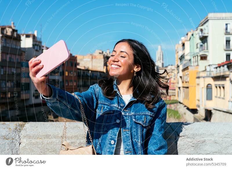 Smiling ethnic woman standing on rooftop and taking selfie smartphone smile using urban building boundary wall young female happy mobile surfing gadget device