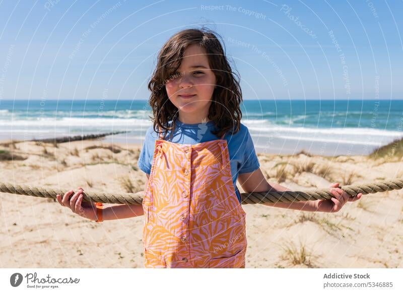 Little girl at rope fence on seashore beach sand smile kid child summer nature happy coast ocean joy water glad resort childhood positive pleasure little