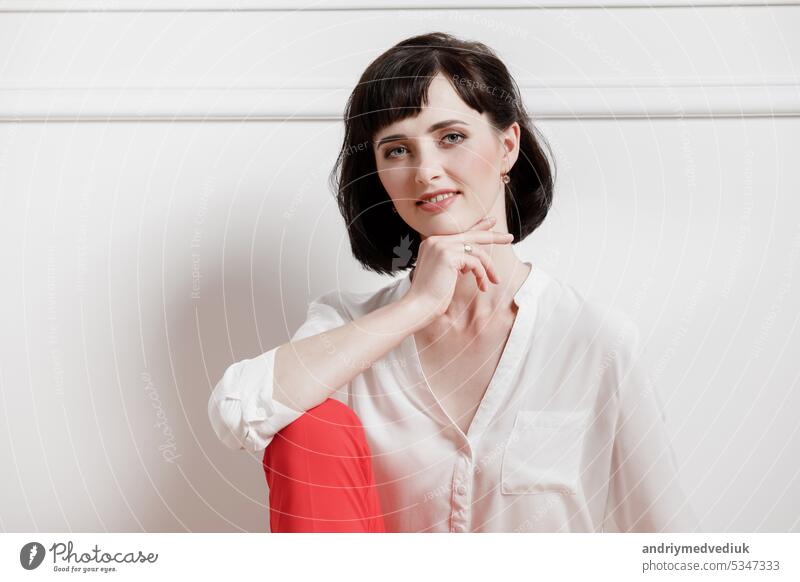 Friendly confident young woman standing with folded arms smiling at the camera over white with copy space. Girl in white shirt and red pants studio background