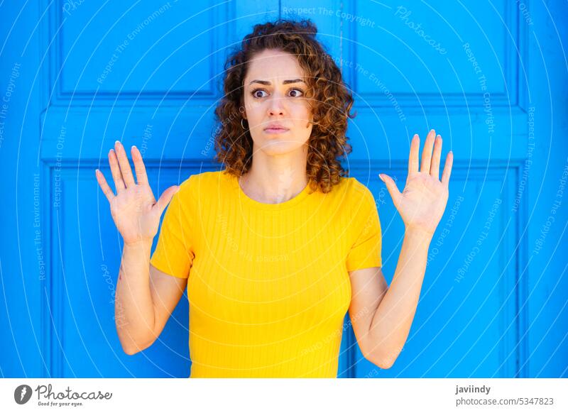 Scared woman with hands up on blue background street afraid scare curly hair pensive city style colorful urban wall thoughtful charming casual female young