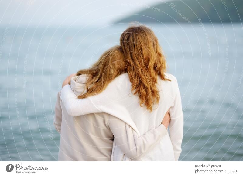 Redhead senior mother and her adult beautiful daughter are walking together on the sea shore. embrace back unrecognizable people maternity family parent