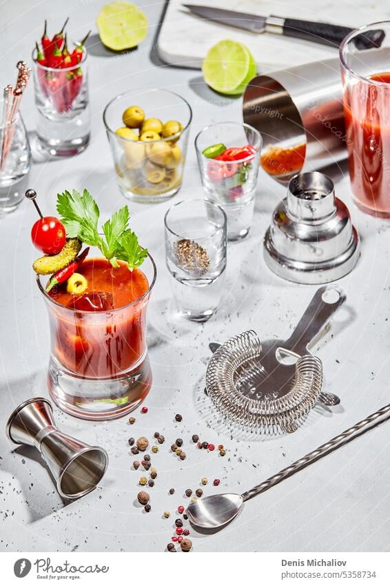 Bloody mary cocktail glass with bar utensils,lime,peppers,olives on light board with sun light. bloody shaker vodka spoon juice strainer tomato cayenne spicy