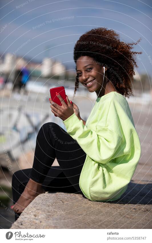 African-American woman sitting by the sea listening to music with her mobile phone in her hand. technology relaxation leisure outdoors beach coastal headphones