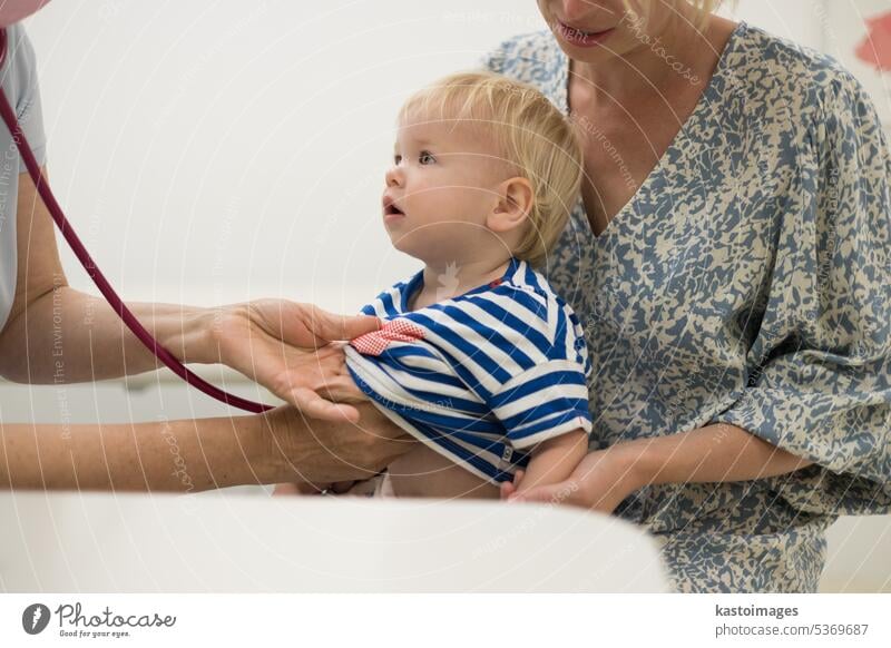 Infant baby boy child being examined by his pediatrician doctor during a standard medical checkup in presence and comfort of his mother. National public health and childs care care koncept.