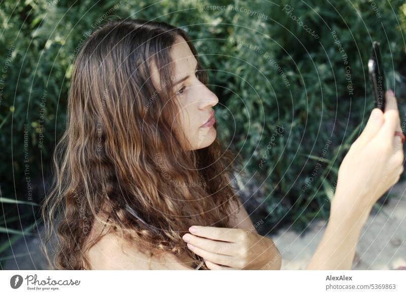 Portrait of young brunette long haired woman looking at her smartphone used as mirror on beach in front of bushy plants Woman Young woman Brunette Long-haired