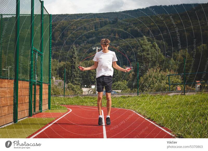 Brown-haired boy with an athletic figure wearing a white T-shirt and black shorts is jumping rope on an athletic oval. Training to improve jumping, coordination and endurance strength