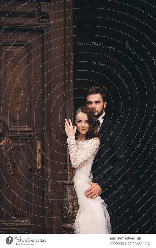 Beautiful newlyweds hugging near the ancient door. Wedding portrait of a stylish groom and a young bride near old house in in a European town embrace beautiful