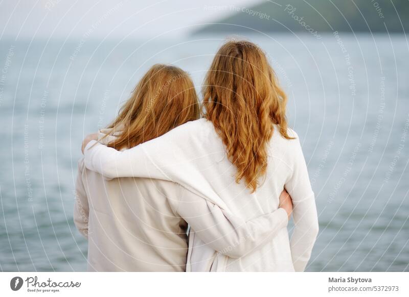 Redhead senior mother and her adult beautiful daughter are walking together on the sea shore. embrace back unrecognizable people maternity family parent