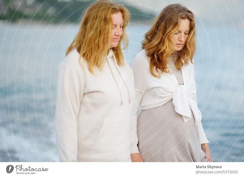 Redhead senior mother and her adult beautiful pregnant daughter are walking together on the sea shore. similar people maternity family look alike talk parent