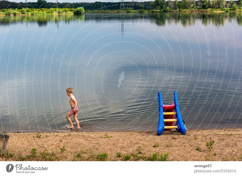 Hesitant - boy walks along the edge of a gravel lake in shallow water gravel pit Swimming lake Bathing place Boy (child) Lake Water Summer Exterior shot