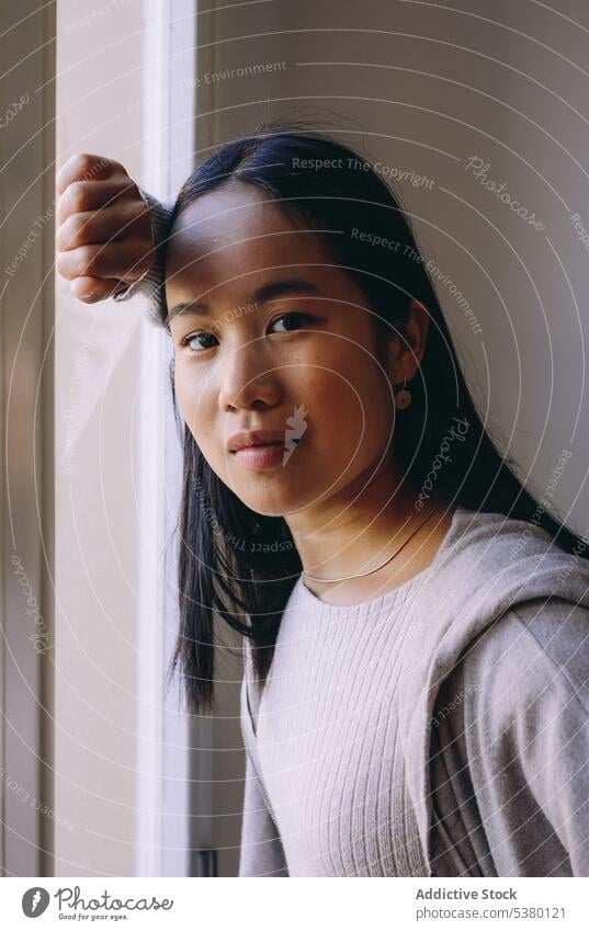 Asian woman looking at camera while standing by window serious portrait confident calm thoughtful pensive style casual female asian woman bright brunette young