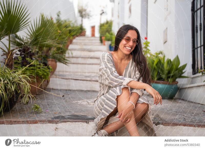 Smiling woman sitting on steps of street smile stair building town plant hand at chin glad positive young female happy cheerful sunglasses potted rest style