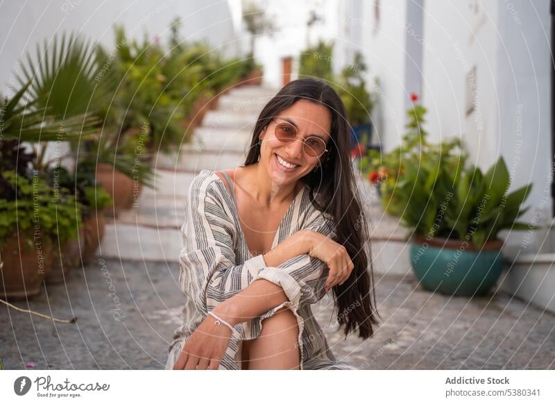 Smiling woman sitting on steps of street smile stair building town plant hand at chin glad positive young female happy cheerful sunglasses potted rest style