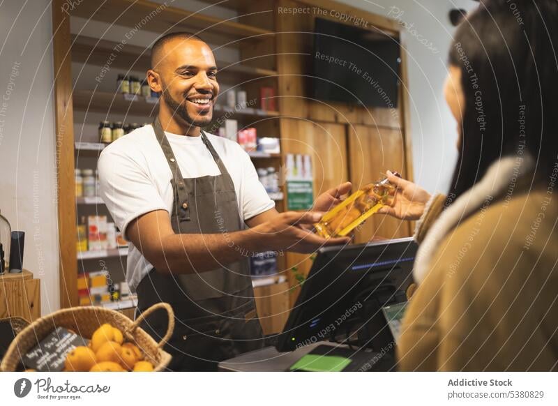 Cheerful black seller giving purchase to customer in shop man woman counter work service owner oil friendly african american positive bottle young smile male