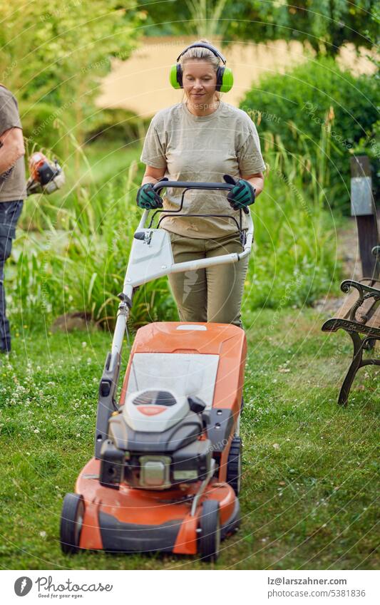 Young Female gardener using a lawn mower while her colleague or bosses is trimming the garden in the background artisan builder chainsaw concept confident