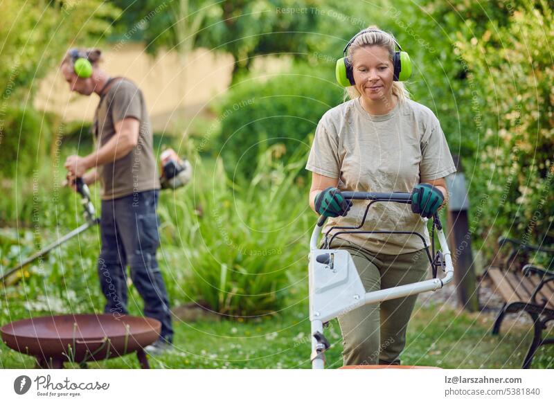 Young Female gardener using a lawn mower while her colleague or bosses is trimming the garden in the background artisan builder chainsaw concept confident