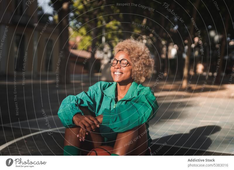 Cheerful black sportswoman resting on sports ground athlete basketball court sportswear cheerful happy sporty sneakers break african american sunset female