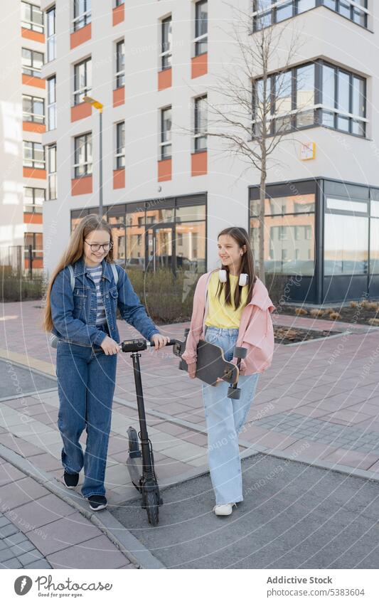 Smiling friends with kick scooter and skateboard walking in city girls smile urban lifestyle leisure together building teenage teenager young sidewalk activity