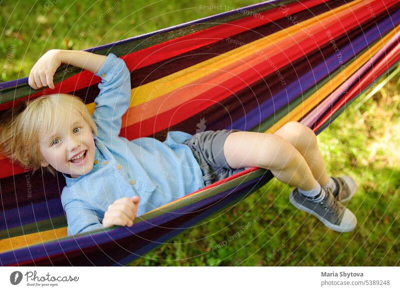 Cute little blond caucasian boy relaxing and having fun in multicolored hammock in backyard or outdoor playground. Summer active leisure for kids. Child swinging on hammock.