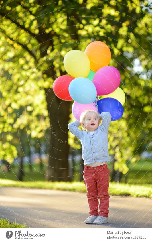 Little boy having fun during celebrating birthday party. Happy child with a bunch of bright multicolor balloons. Preschoolers or toddlers baby birthday party in sunny park. Summer outdoor festival