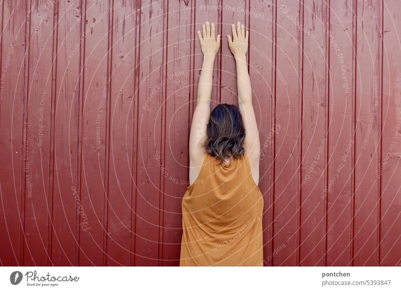 back view of a woman in a yellow summer dress. the woman stands close to a red board wall and presses her hands against it with her arms stretched up. Woman