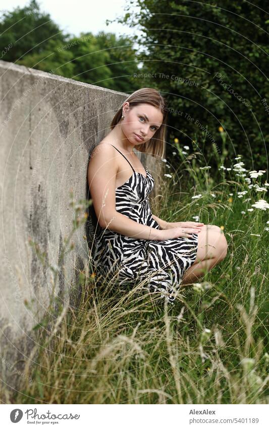 Full body portrait of young woman with makeup on, wearing black and white striped summer dress, leaning against concrete wall on outdoor lawn Woman Young woman