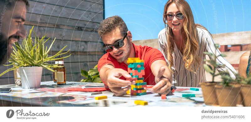 Concentrated man catching jenga game piece next to colleagues on terrace happy young playing pushing block smile concentrated friends teamwork worker break